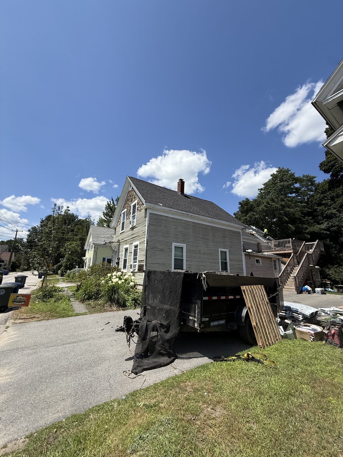 Black dump trailer parked in a driveway to collect tear off debris beside a grey multi family house, with new shingle bundles and materials staged on the lawn